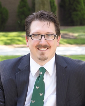 Headshot of Paul taken outside in nature. He is wearing a blue suit, a green tie, and glasses