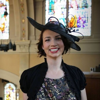 Jen in a church in semi-formal clothing with a jaunty black hat on, smiling